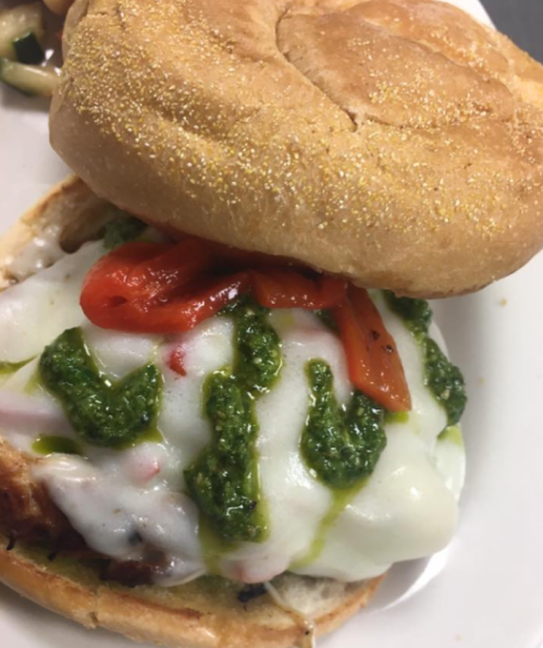 A close-up of a sandwich with a toasted bun, grilled chicken, melted cheese, roasted red peppers, and pesto drizzle.