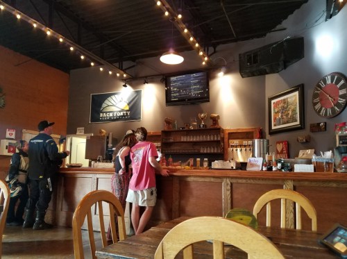 A cozy bar interior with patrons at the counter, rustic decor, and warm lighting. A TV displays information on the wall.
