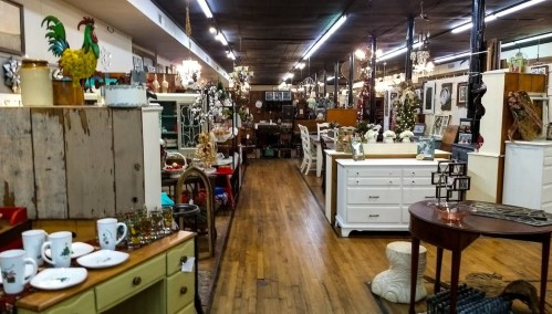 A spacious antique shop filled with vintage furniture, decor, and collectibles, featuring wooden floors and bright lighting.