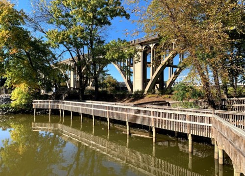 A wooden walkway beside a calm waterway, with a bridge and trees in the background under a clear blue sky.
