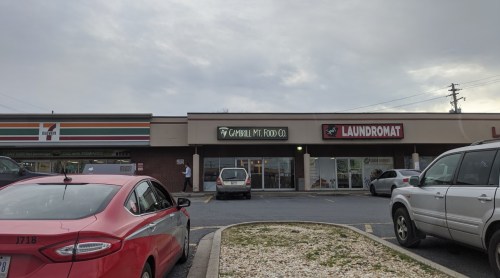 A shopping plaza featuring a laundromat and a food company, with parked cars in the foreground and cloudy skies above.