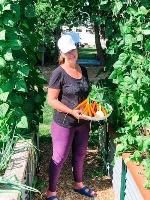 A woman in a garden holds a plate of freshly harvested carrots and vegetables, surrounded by green plants.