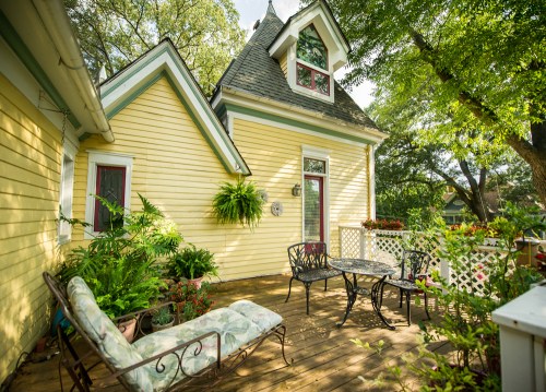 A charming yellow house with a cozy porch, featuring a seating area surrounded by lush greenery and potted plants.