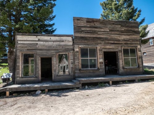 A weathered wooden building with large windows, surrounded by trees and a clear blue sky.