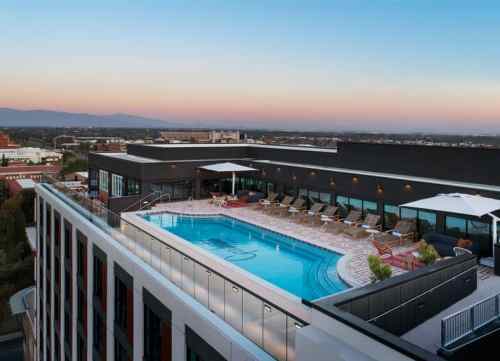 Rooftop pool with lounge chairs and umbrellas, overlooking a city skyline at sunset.