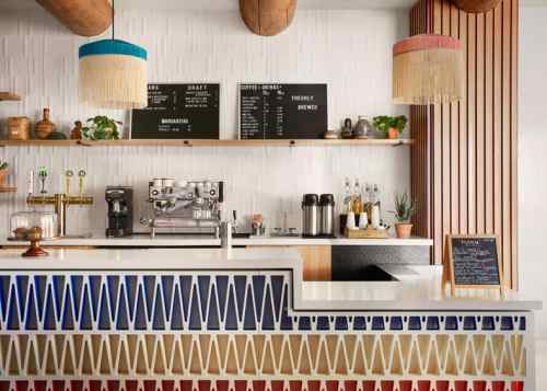 A modern café bar with a colorful patterned counter, coffee machine, and menu boards, featuring hanging light fixtures.