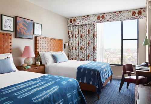 A stylish hotel room with two beds, colorful bedding, a desk, and large windows letting in natural light.