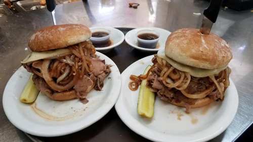 Two hearty sandwiches with roast beef, melted cheese, and sautéed onions, served with pickles and dipping sauces.