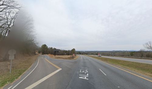 A fork in the road with a sign for AL-53 X, surrounded by trees and a cloudy sky in the background.