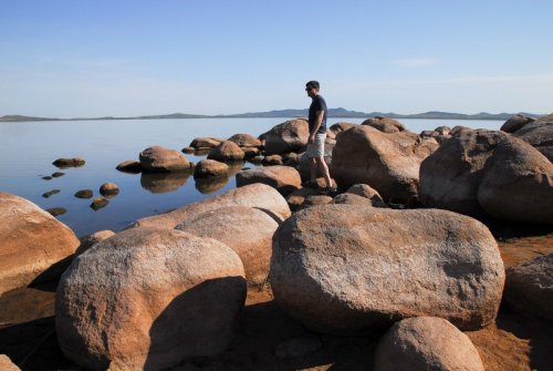 A person stands on large rocks by a calm lake, with distant mountains under a clear blue sky.