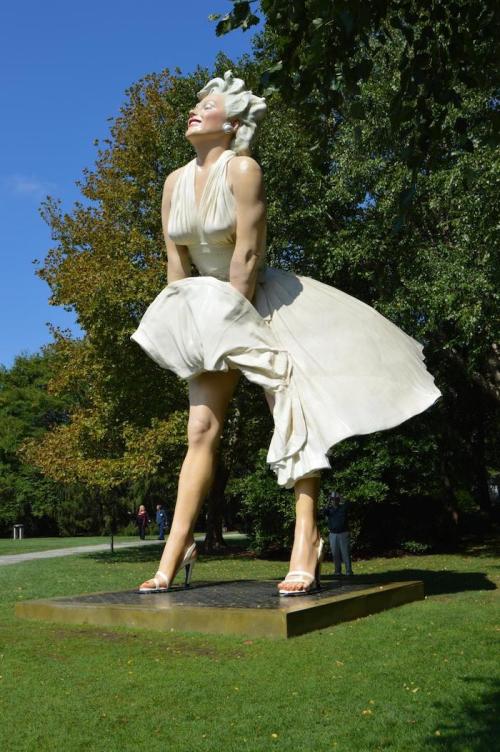 A large statue of a woman in a white dress, smiling with her skirt blown up, set in a green park.