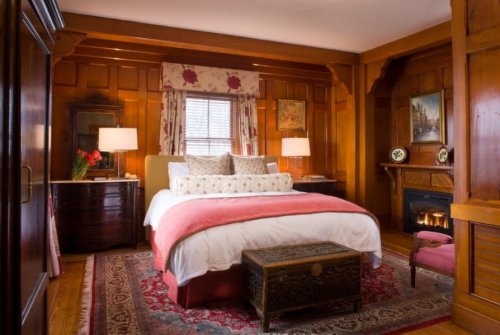 Cozy bedroom with wooden walls, a large bed, decorative pillows, a fireplace, and elegant furnishings.