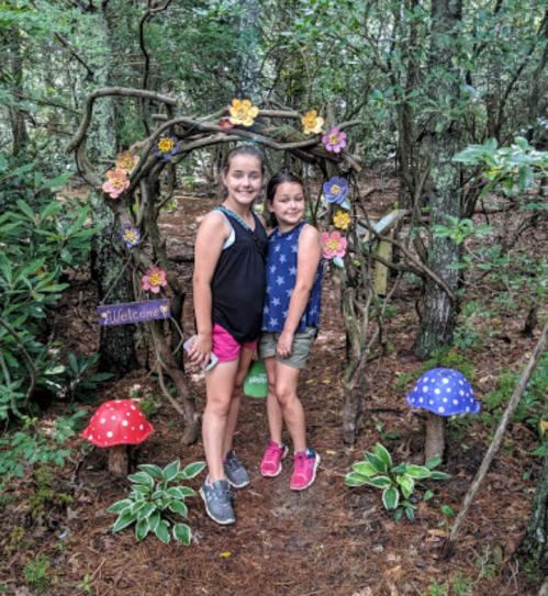 Two girls stand under a whimsical arch adorned with flowers and mushrooms in a forest setting.