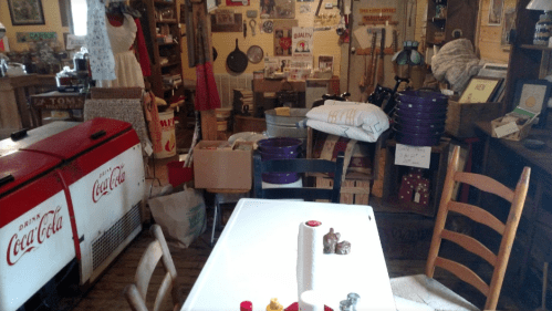 A cluttered vintage room filled with memorabilia, old furniture, and Coca-Cola coolers, creating a nostalgic atmosphere.