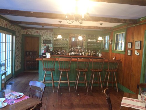 Cozy bar area with wooden stools, green walls, and a variety of bottles on shelves, surrounded by floral wallpaper.