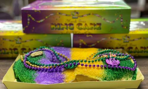 A colorful king cake with purple, green, and gold sprinkles, adorned with beads, in front of a festive box.