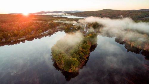 Aerial view of a serene lake at sunrise, with mist rising over trees and colorful autumn foliage reflecting in the water.