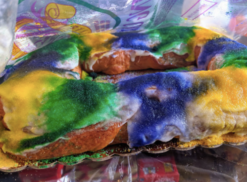 A colorful King Cake decorated with green, yellow, and purple sugar, displayed in a clear plastic bag.