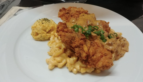 A plate featuring fried chicken on creamy macaroni and cheese, with a side of cornbread and green onion garnish.