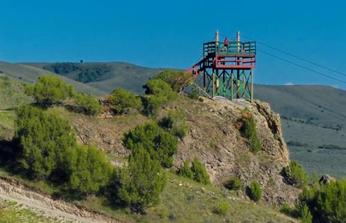A colorful observation tower on a rocky hill, surrounded by green shrubs and mountains under a clear blue sky.