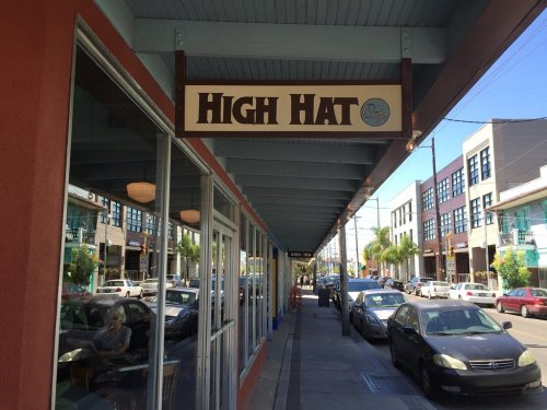 Sign for "High Hat" cafe under a blue awning, with a view of a street lined with parked cars and buildings.