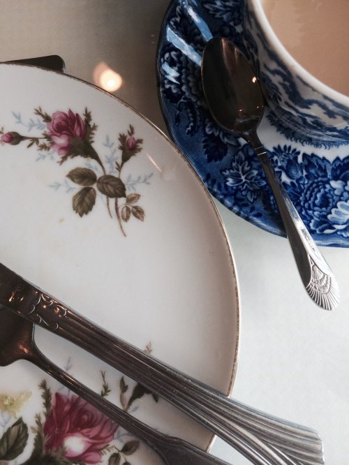 A close-up of a floral-patterned plate, silver cutlery, and a blue and white teacup on a glass table.