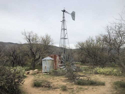 A weathered windmill stands near a rusted water tank in a desert landscape with sparse vegetation and distant mountains.