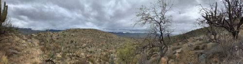 A panoramic view of a desert landscape with rocky terrain, sparse vegetation, and a cloudy sky.