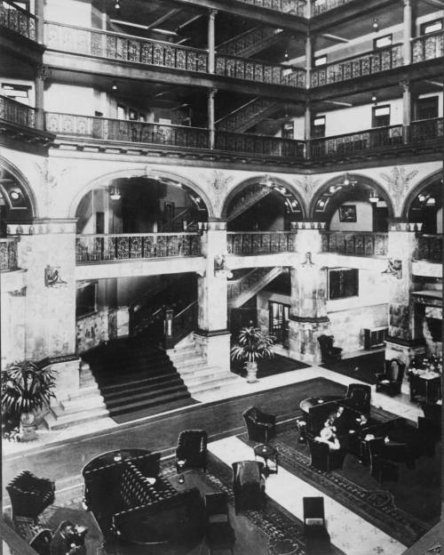 Historic hotel lobby featuring ornate architecture, grand staircase, and elegant furnishings. Black and white photo.