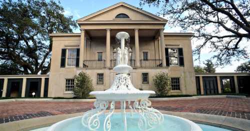 A grand, two-story mansion with columns, featuring a white fountain in the foreground and lush greenery around.