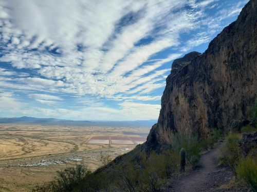 A scenic view of a rocky landscape with a winding path, expansive sky, and distant fields under a blue and cloudy sky.