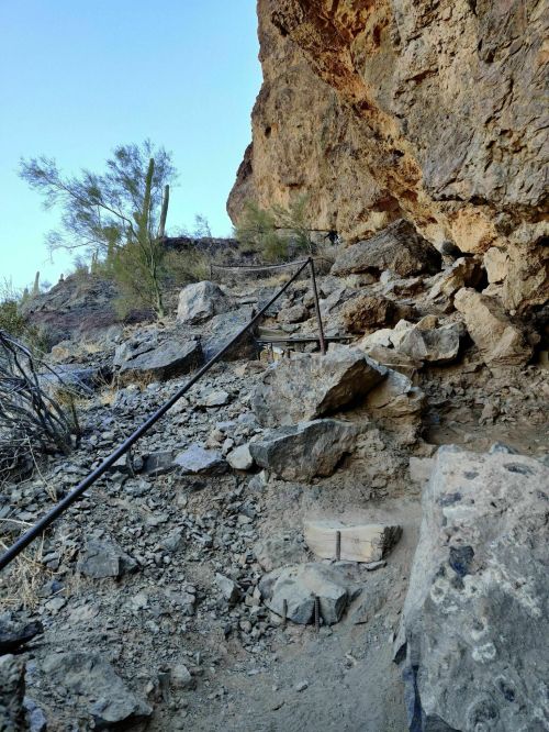 Rocky trail with a metal railing leading up a steep incline, surrounded by desert vegetation and rocky terrain.