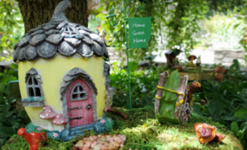 A whimsical fairy house with a mushroom garden and a "Home Sweet Home" sign surrounded by greenery.