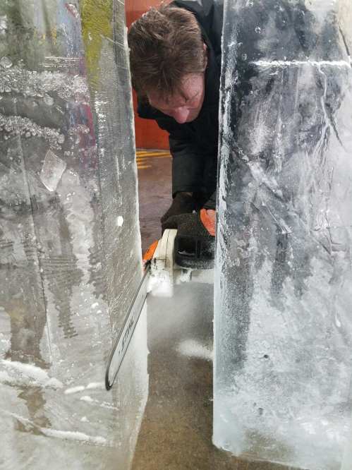 A person using a chainsaw to cut through large blocks of ice in a workshop setting.
