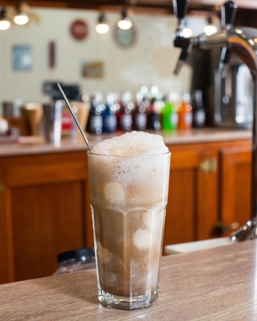 A frothy glass of iced coffee with ice cubes, set on a wooden counter with colorful drinks in the background.