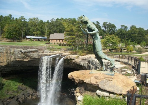 A bronze statue of a Native American stands near a waterfall, surrounded by lush greenery and a rustic building in the background.
