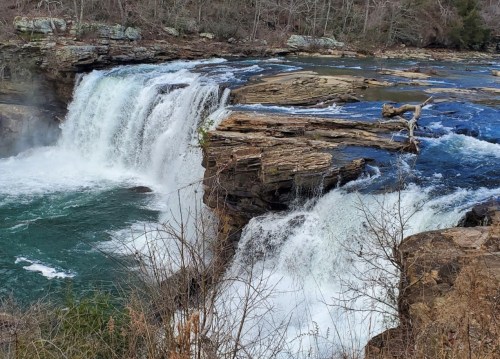 A scenic view of a waterfall cascading over rocky cliffs into a turquoise pool, surrounded by trees and natural landscape.