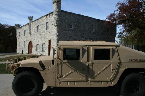 A tan military Humvee parked in front of a stone building with castle-like features and a clear blue sky.