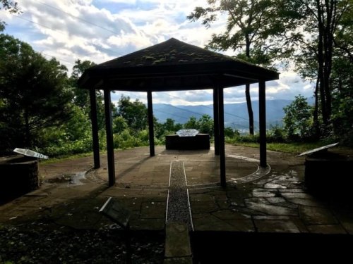 A gazebo with a stone floor overlooks a scenic mountain view, surrounded by trees and cloudy skies.