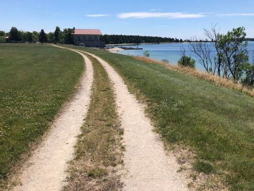 A sandy path leads along a grassy area by a calm lake, with trees and a building in the background under a clear blue sky.