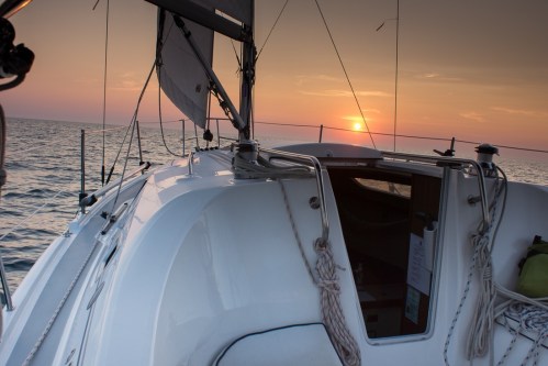 A sailboat deck at sunset, with the sun setting over calm waters and a clear sky.