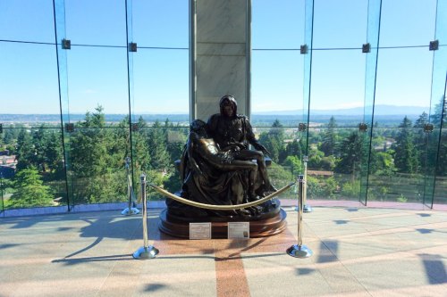 A bronze statue of a woman holding a man, set against a backdrop of trees and mountains, inside a glass building.