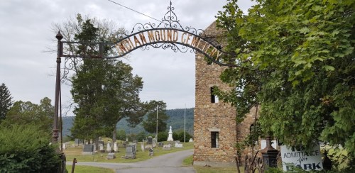 Entrance to Indian Mound Cemetery, featuring a stone archway and gravestones, set against a cloudy sky.
