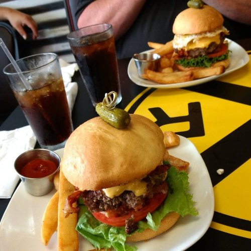 A close-up of a cheeseburger with lettuce, tomato, and a pickle, served with fries and drinks in a diner setting.