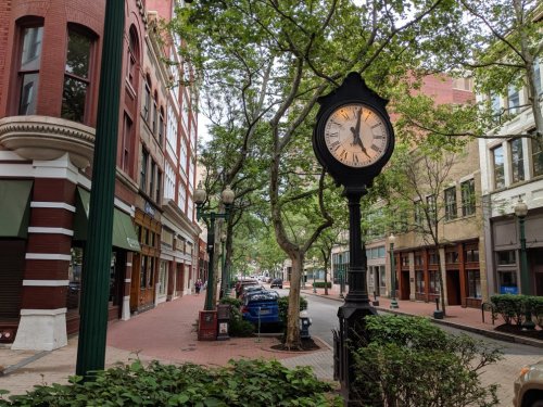 A street view featuring a vintage clock, trees, and brick buildings, with parked cars along the sidewalk.