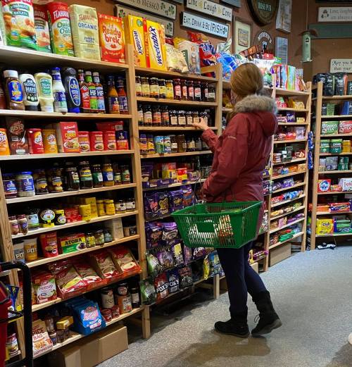 A person shopping in a grocery store aisle filled with various food products and spices, holding a green basket.