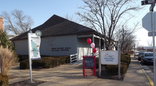 A welcome center building with signs, balloons, and a chalkboard menu outside, surrounded by shrubs and trees.