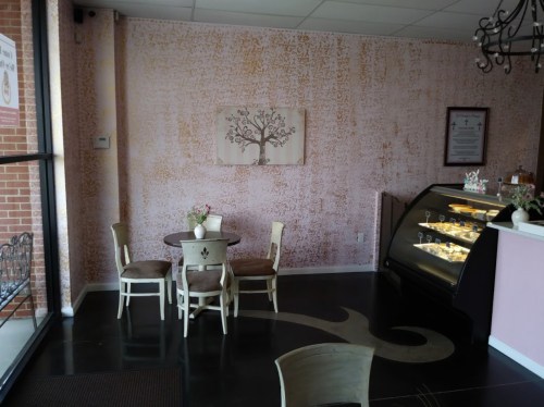 Cozy café interior with a round table, chairs, a display case of pastries, and a decorative tree painting on the wall.