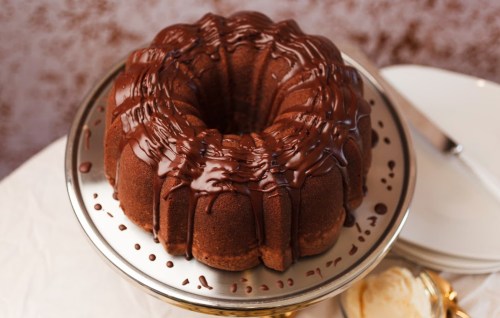 A chocolate bundt cake drizzled with chocolate ganache, displayed on a silver cake stand.