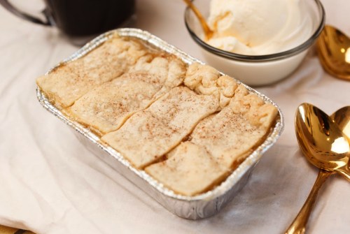 A silver foil dish of baked dessert with a golden crust, served alongside a bowl of vanilla ice cream and golden spoons.